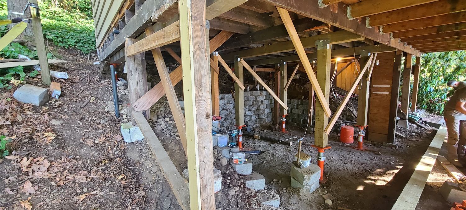 Wooden structure being repaired, supported by jacks and bracing. Construction site outdoors, dirt ground.