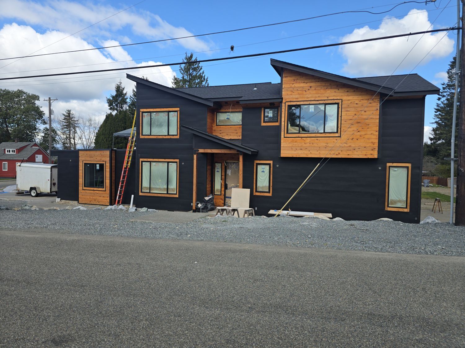 Modern two-story house with black siding and wood accents, under construction, on a gravel lot under a blue sky.