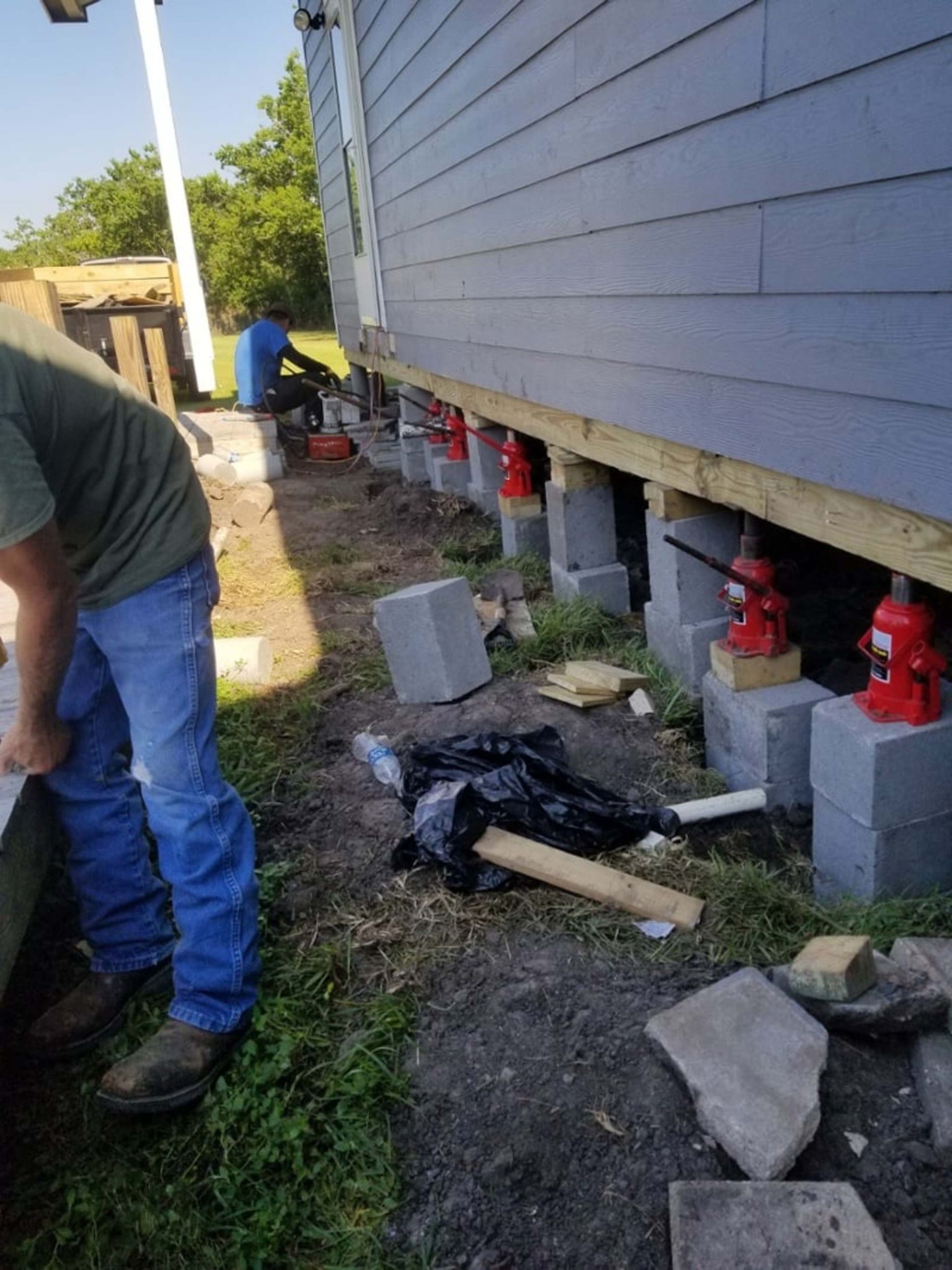 House foundation being repaired with jacks and cinder blocks. Two people work outside the blue-sided building.