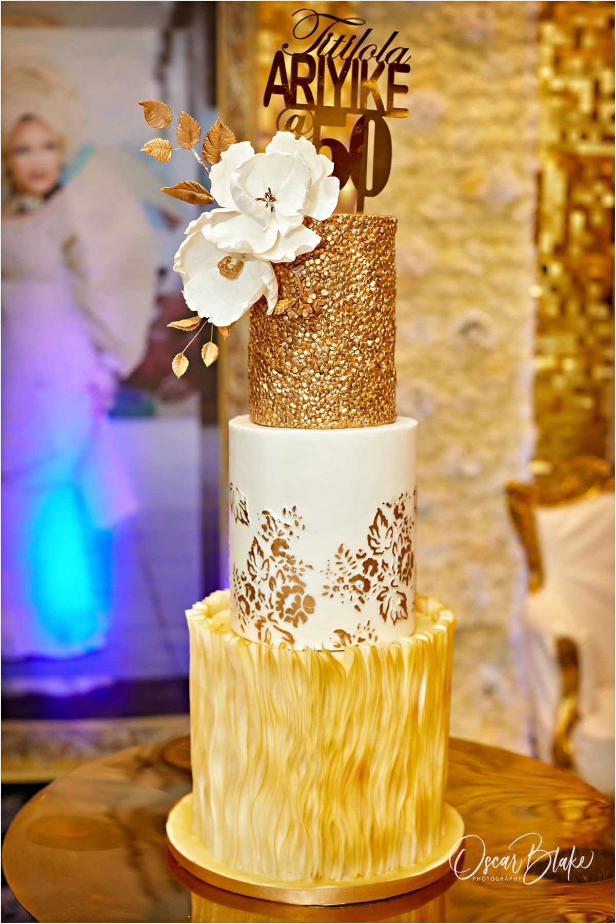 Three-tiered gold and white birthday cake with gold accents, flowers, and a 