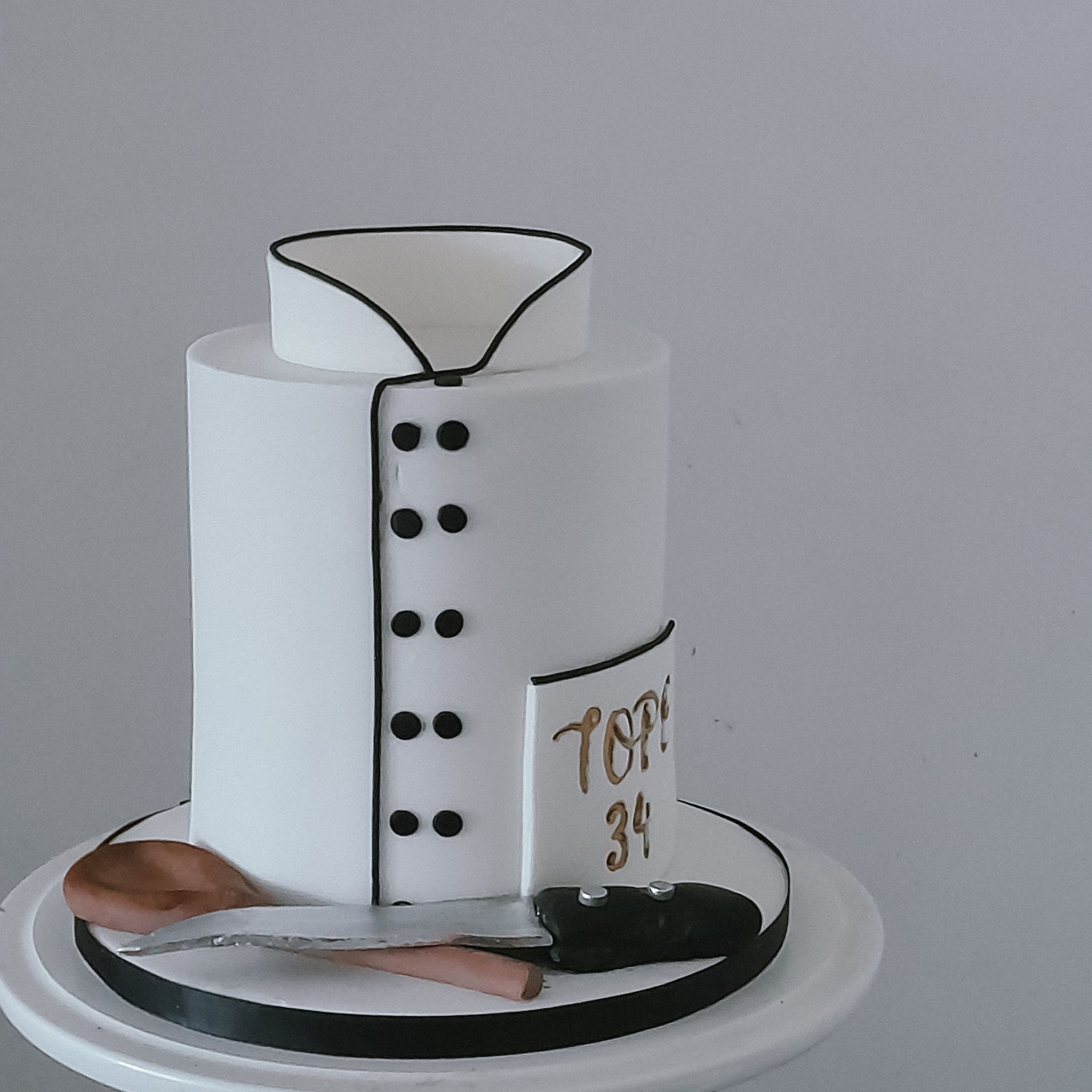 Cake decorated as a chef's jacket, with black details and a wooden spoon and knife at the base, says 