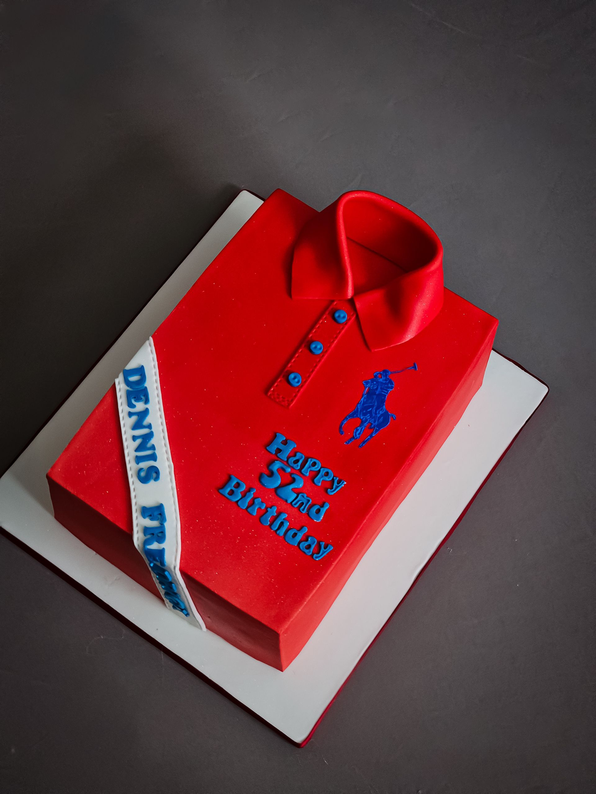 Red polo shirt cake with blue accents, white ribbon with text, and text wishing 
