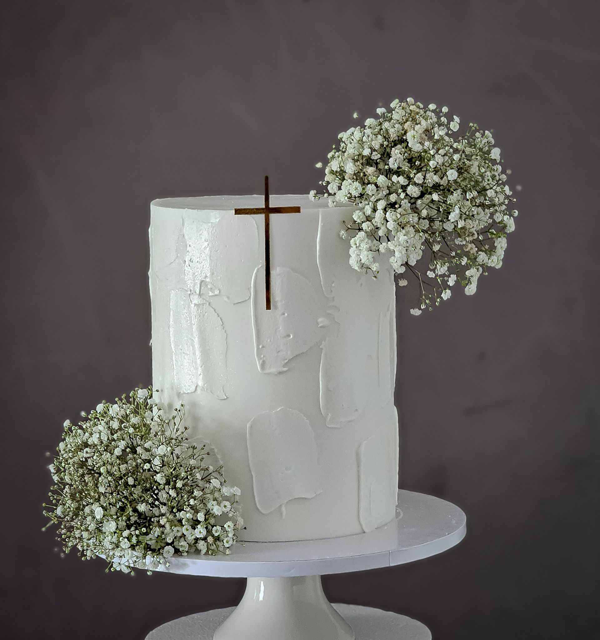 White frosted cake on a pedestal, decorated with a gold cross and two clusters of white baby's breath.
