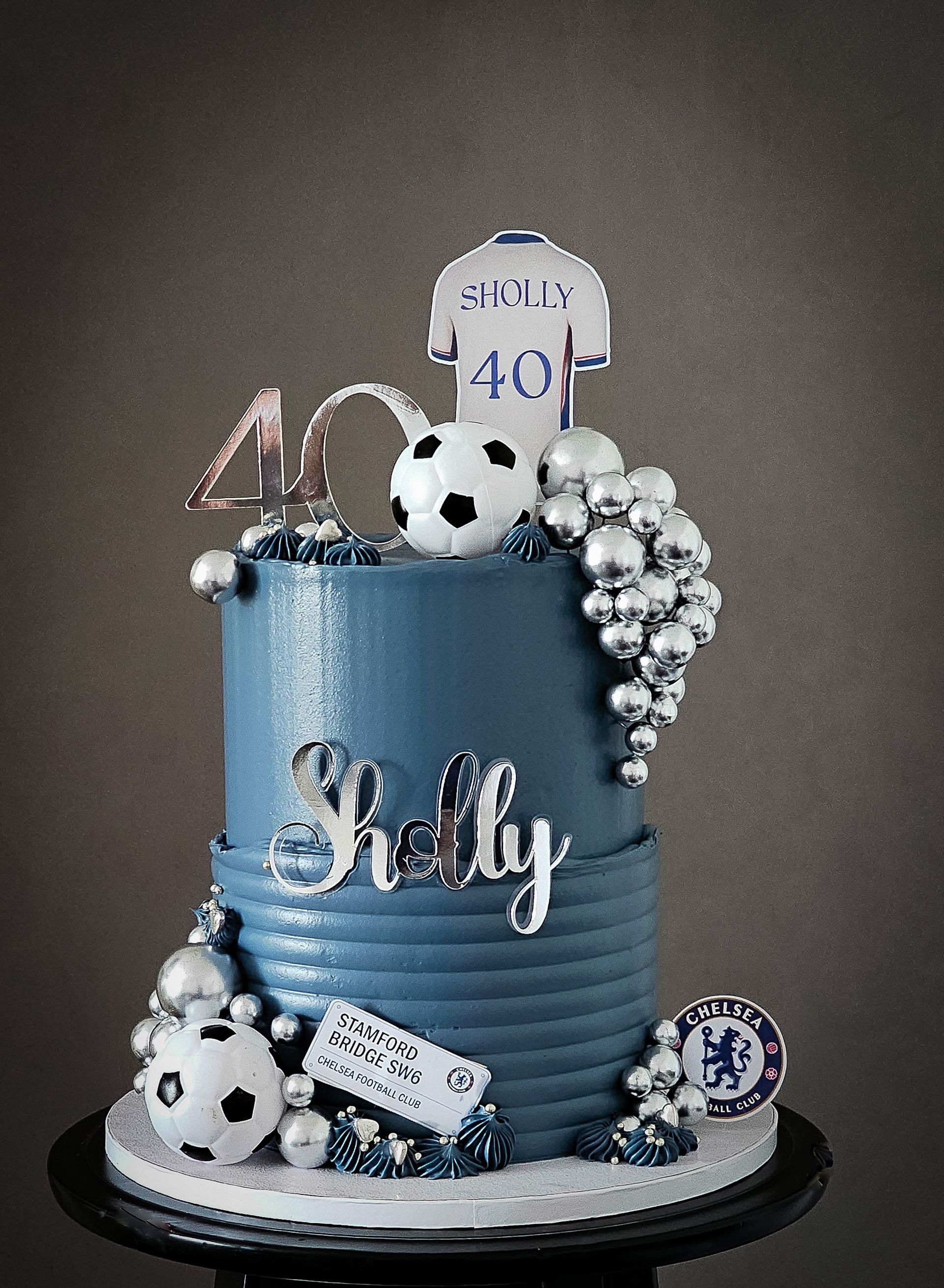 Blue tiered birthday cake with soccer theme, silver accents, a Chelsea jersey topper, and the name 