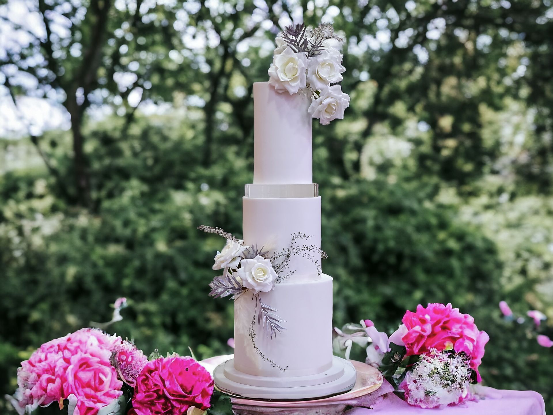 Four-tiered white wedding cake decorated with white roses and greenery, placed outdoors with pink flowers in the foreground.