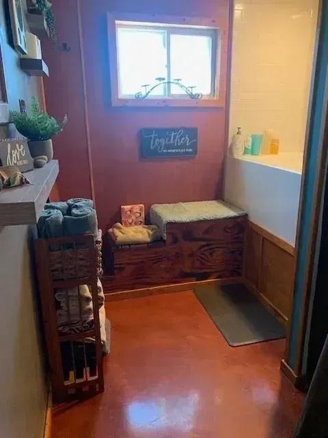 Cozy bathroom with red floors, a wooden bench, a window, and a shower/tub combo. A wooden shelf holds plants and decor.