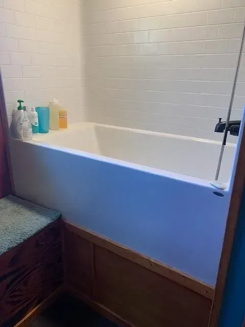 White bathtub with white tiled wall, wooden trim, and bottles on the edge.