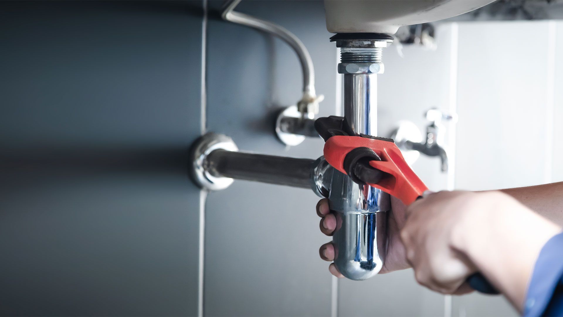 Hands of a plumber working on pipes under a sink; chrome fixtures and gray tiled wall.