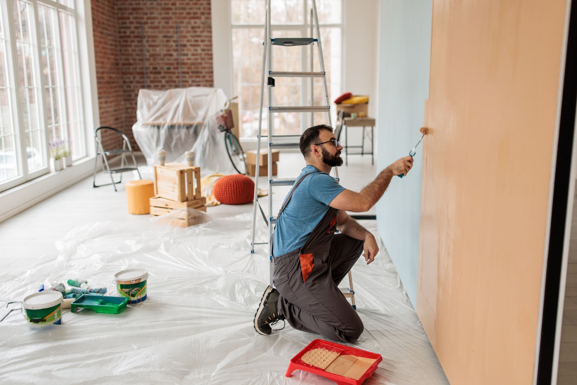 Person kneeling while painting a wall with a roller in a bright, prepped living space.