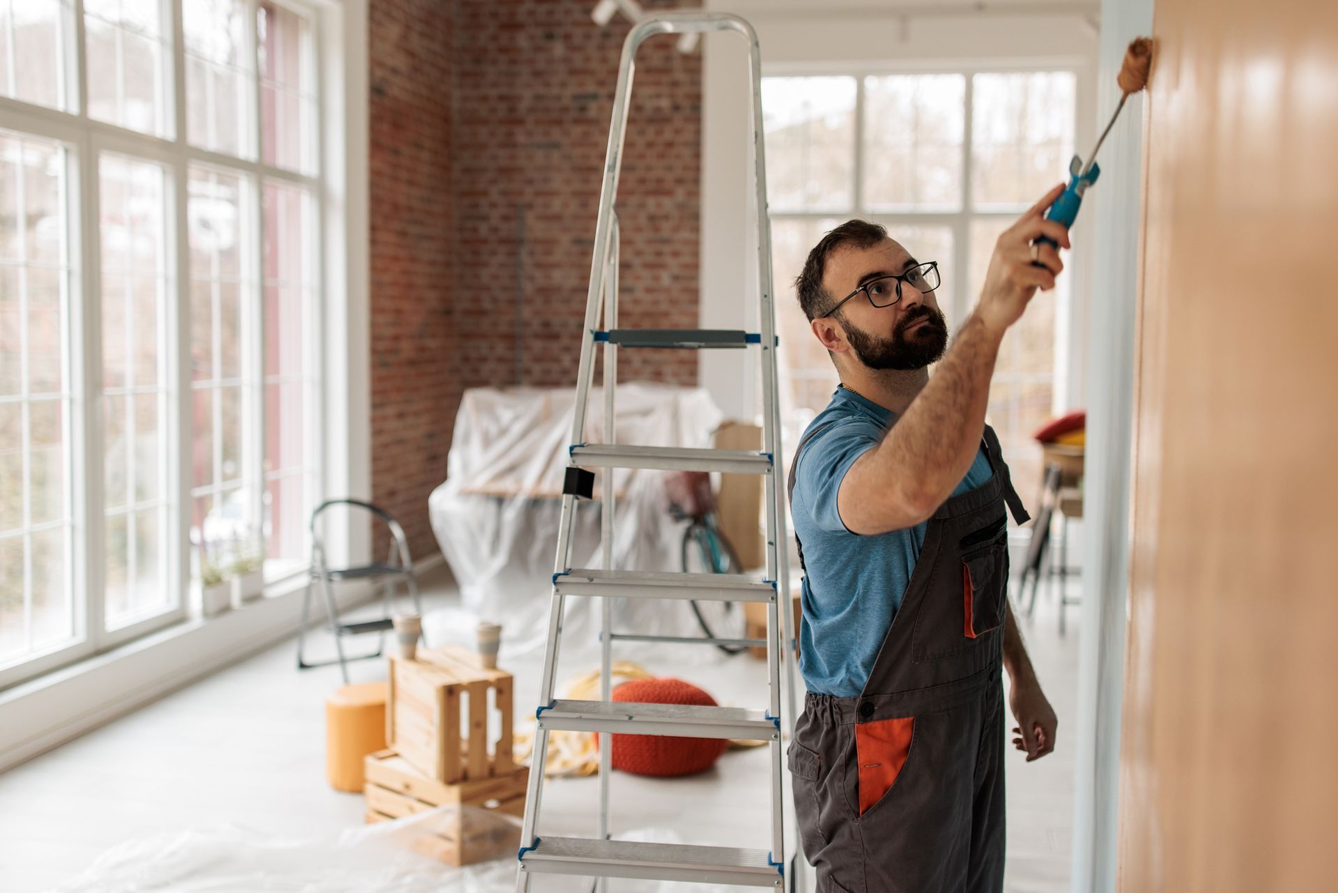 An interior painter, wearing a blue shirt and dark overalls, is applying paint on wall surface.