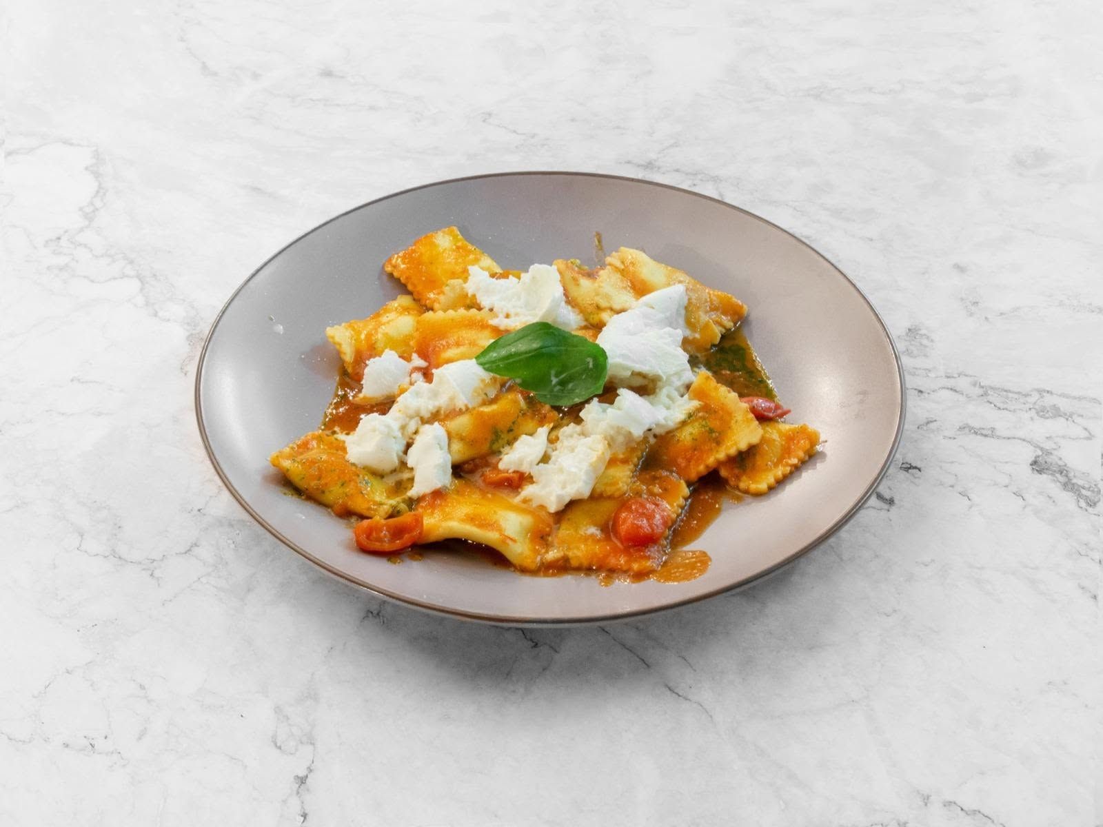 Pasta su un piatto grigio condita con besciamella e basilico su una superficie di marmo chiaro.
