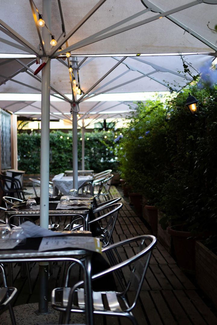 Tavolini all'aperto del caffè sotto ombrelloni bianchi, accanto a una siepe e a una terrazza in legno.