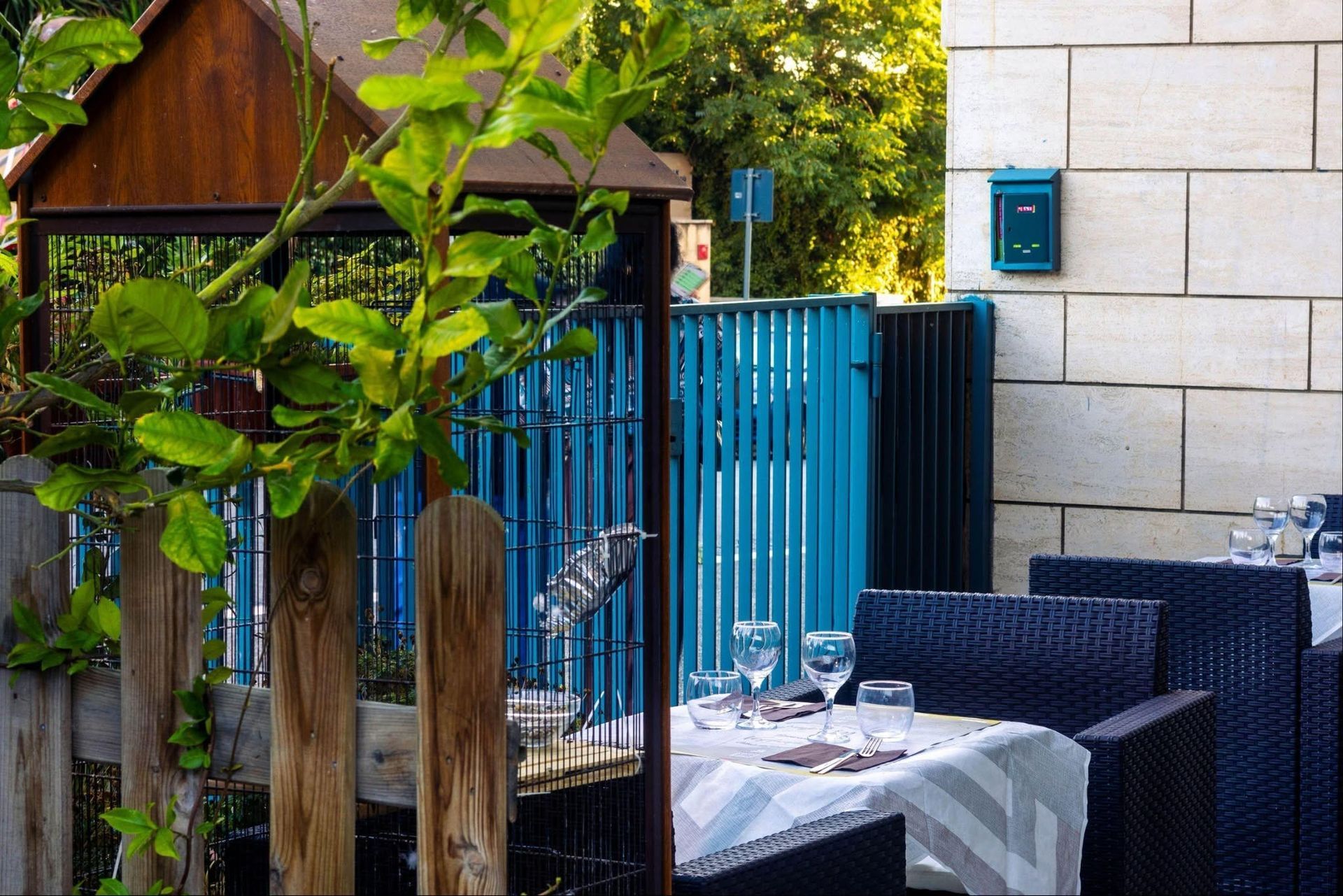 Patio esterno recintato di blu con tavoli, sedie e piante, accanto a una casa.