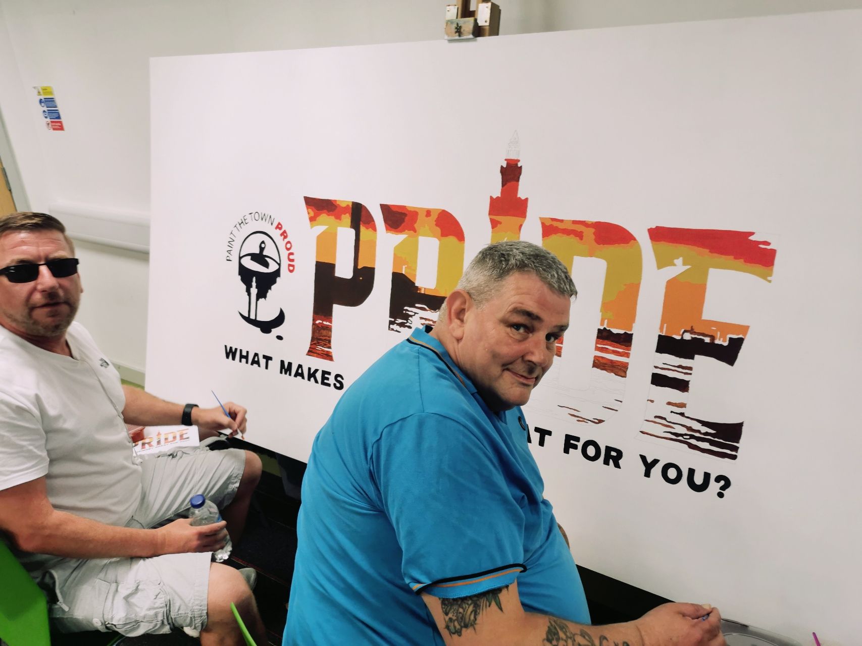 Two men are sitting in front of a sign that says pride