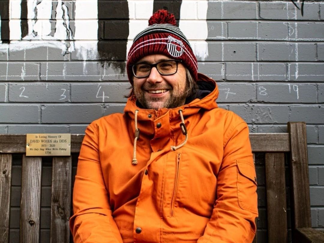 A man wearing a hat and glasses is sitting on a bench in front of a brick wall.