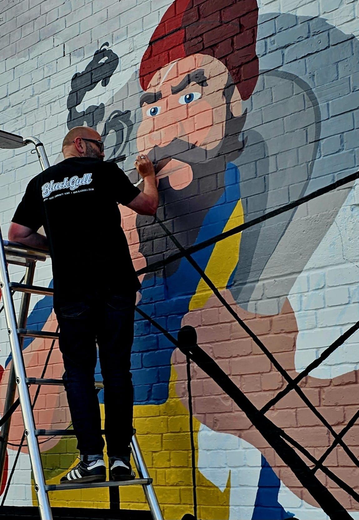 A man is standing on a ladder painting a mural on a brick wall.