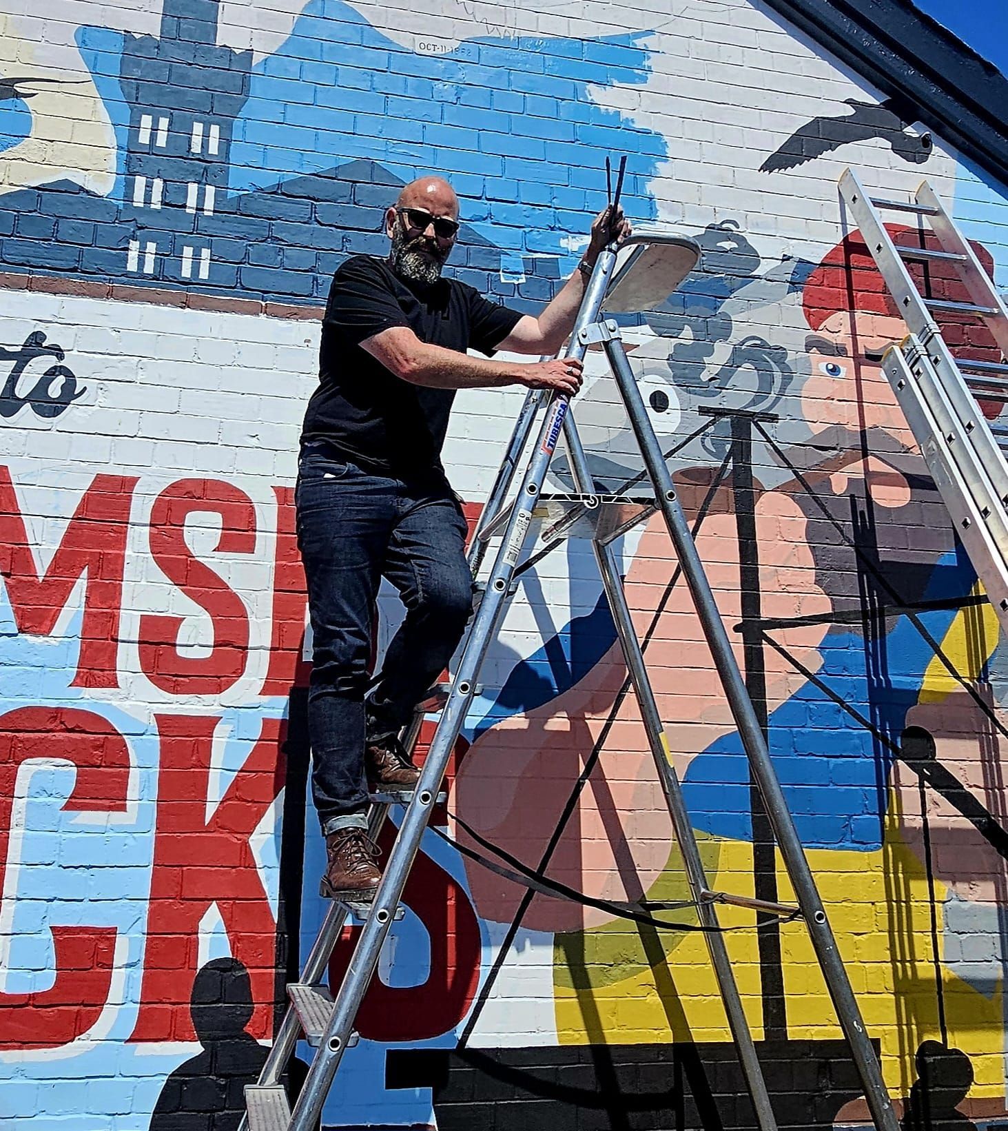 A man is standing on a ladder painting a mural on a brick wall.