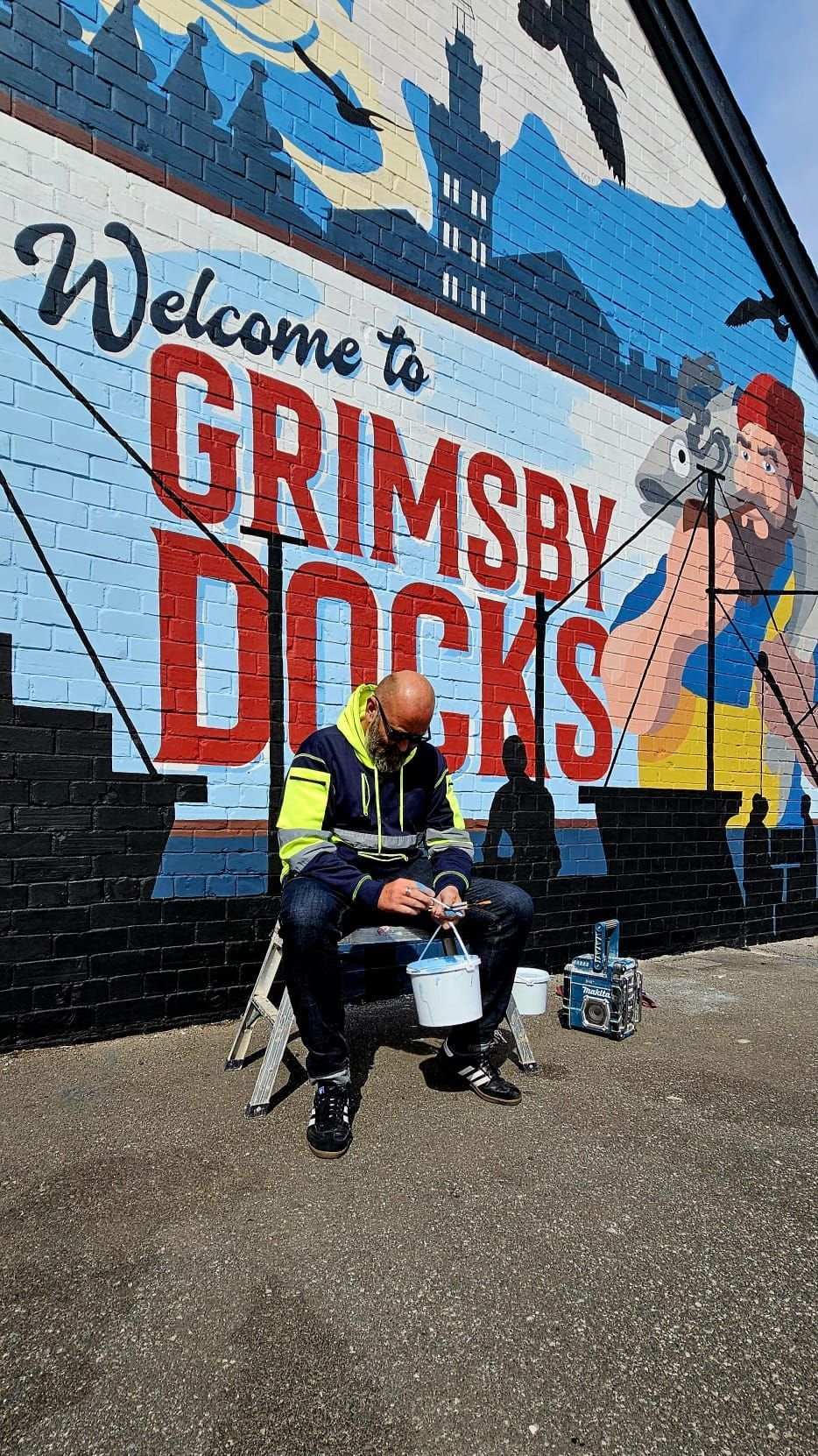 A man is sitting in front of a mural that says welcome to grimsby docks.