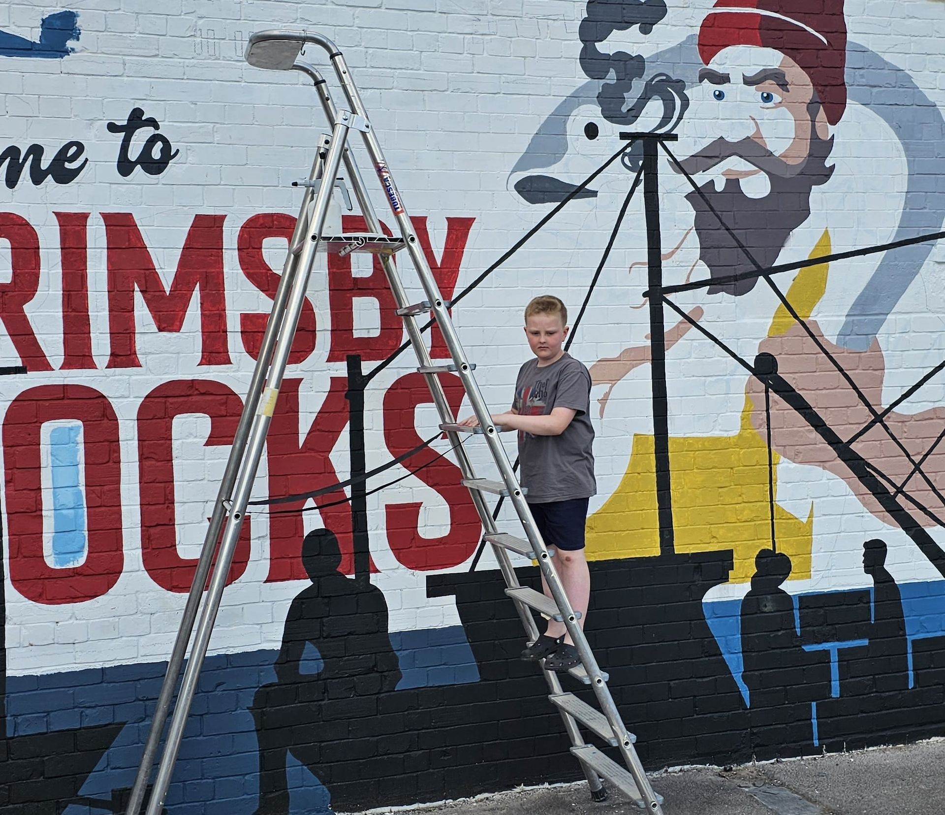 A boy is standing on a ladder in front of a mural that says welcome to rimsby docks