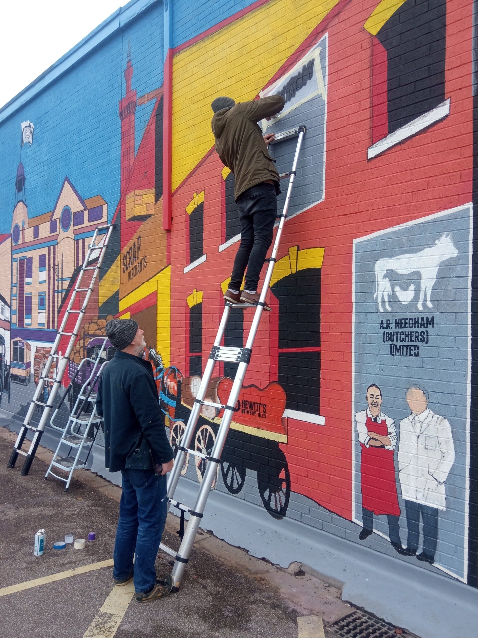 A man on a ladder is painting a mural on the side of a building.
