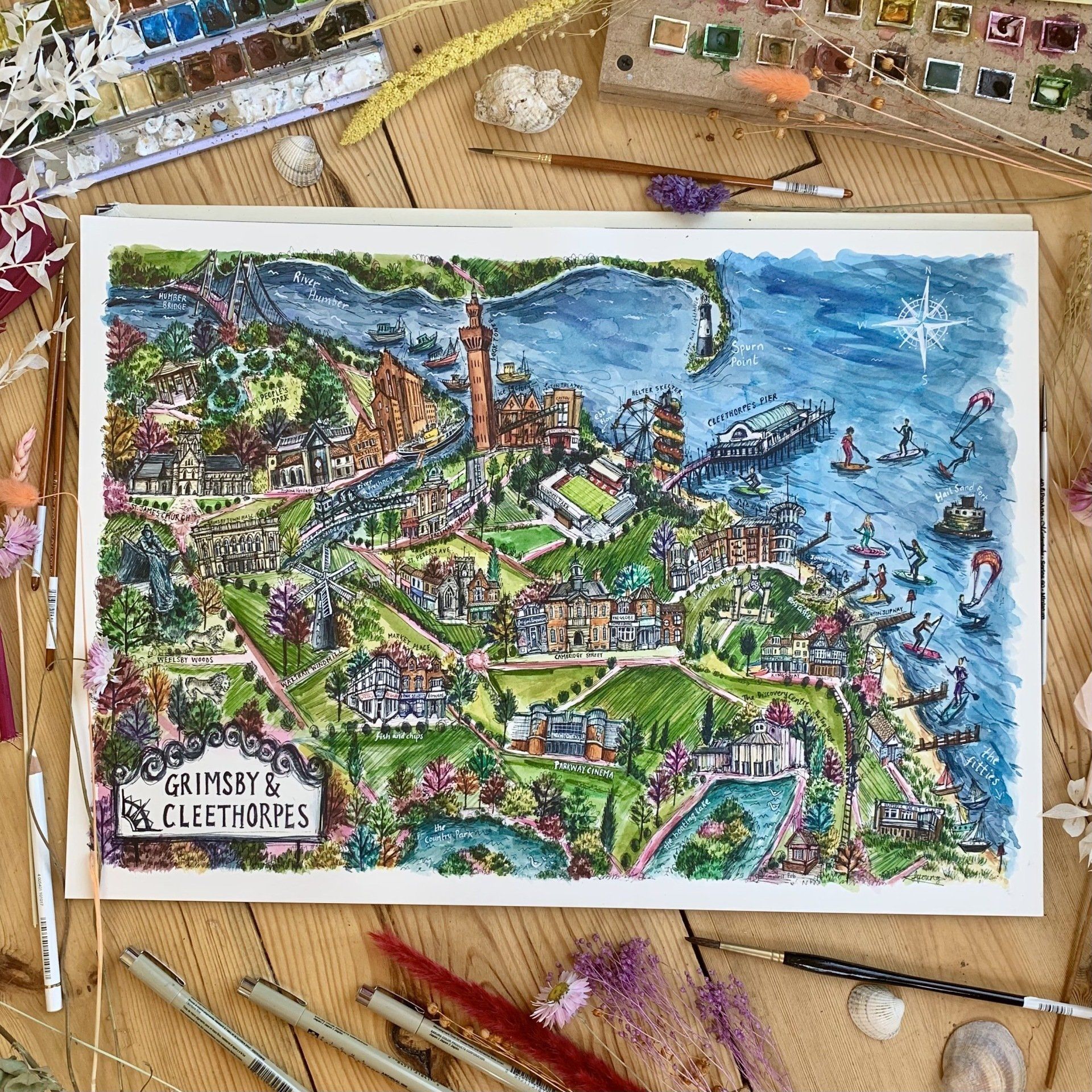 A painting of a city is sitting on a wooden table surrounded by brushes and paints.