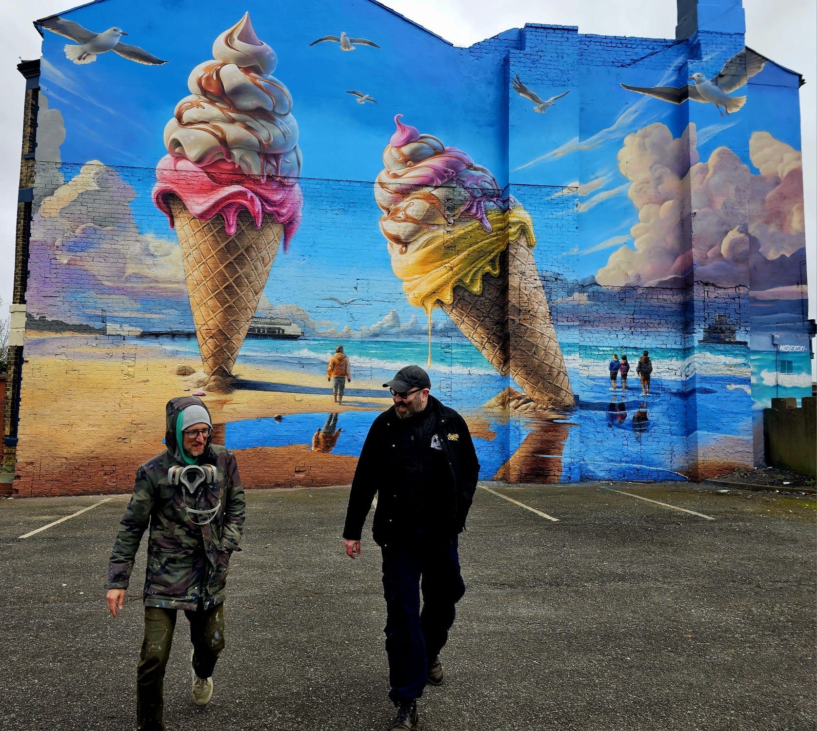 Two men are walking in front of a large mural of ice cream cones on the side of a building