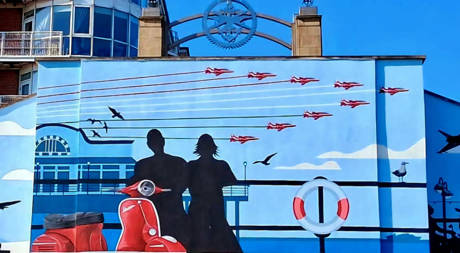A mural on the side of a building shows a man and woman on a scooter