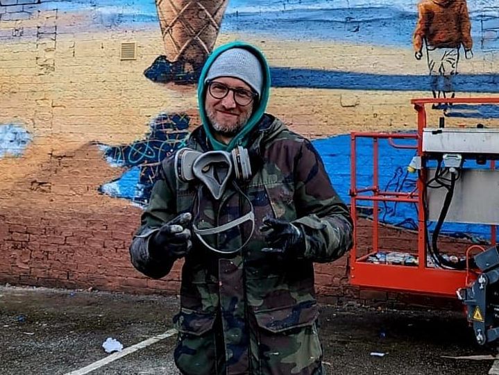 A man wearing headphones and a mask is standing in front of a mural.