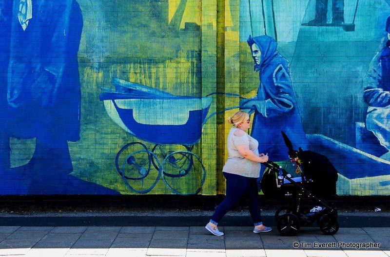 A woman pushing a stroller down a sidewalk in front of a mural.