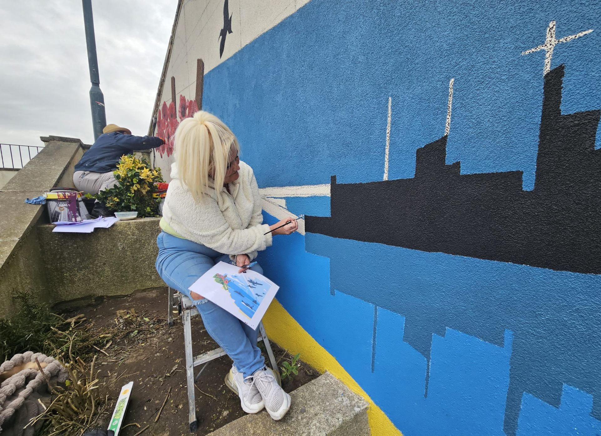 A woman is sitting on a ladder painting a mural on a wall.
