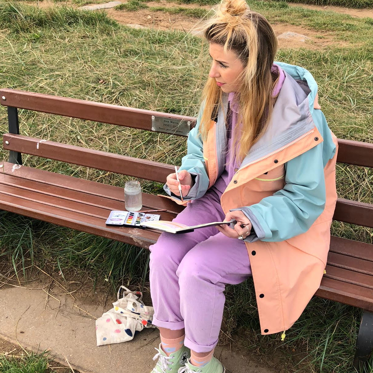 A woman is sitting on a park bench holding a book and a pen.