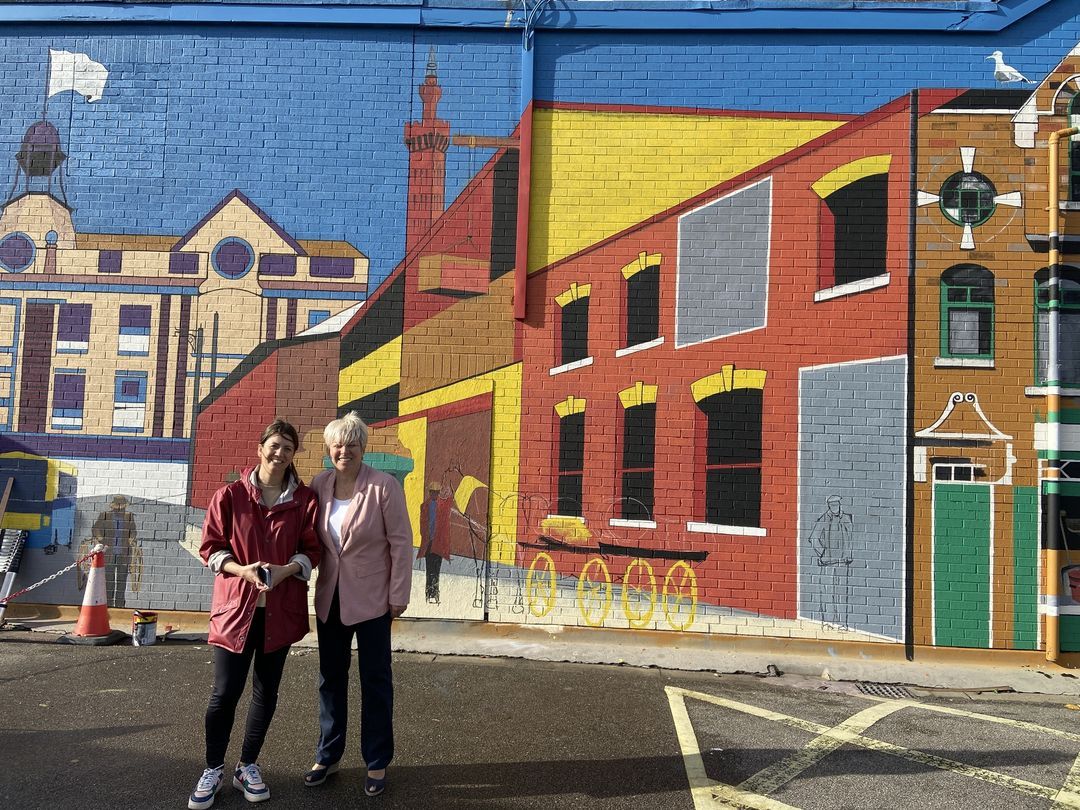 Two women are standing in front of a large mural on the side of a building.