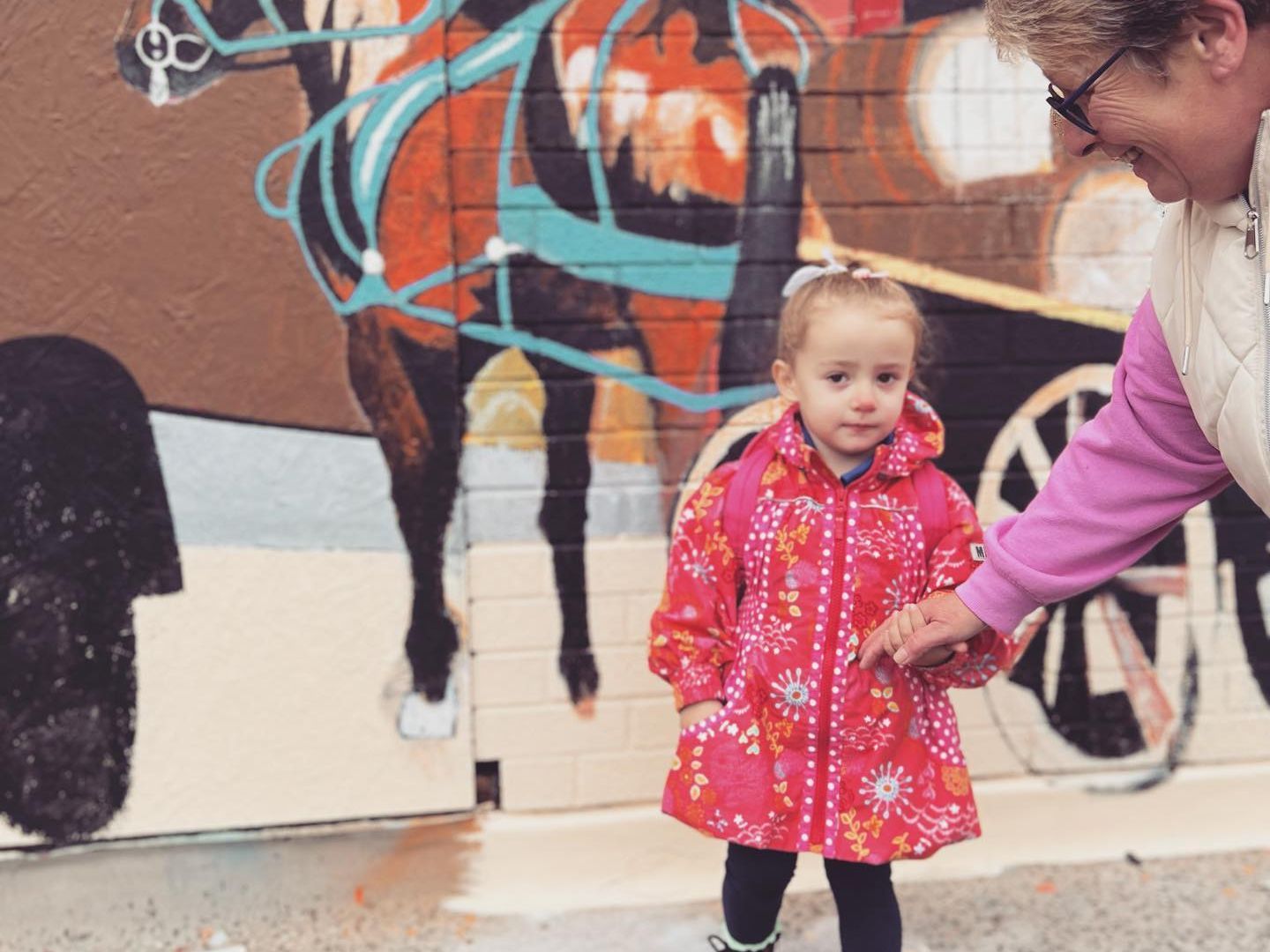 A little girl standing next to a woman in front of a horse drawn carriage mural
