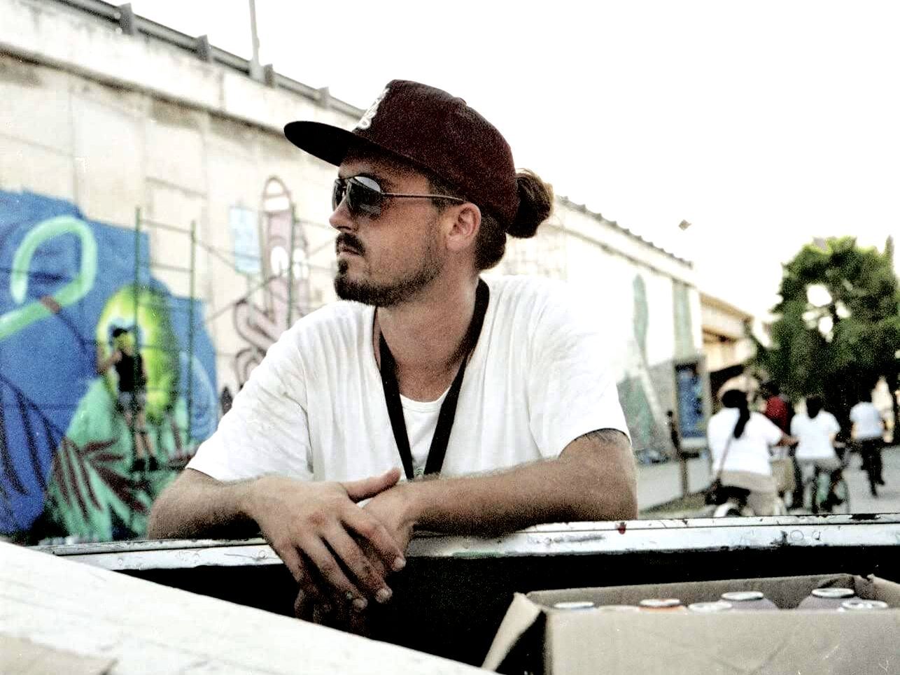 A man wearing a hat and sunglasses leans against a wall