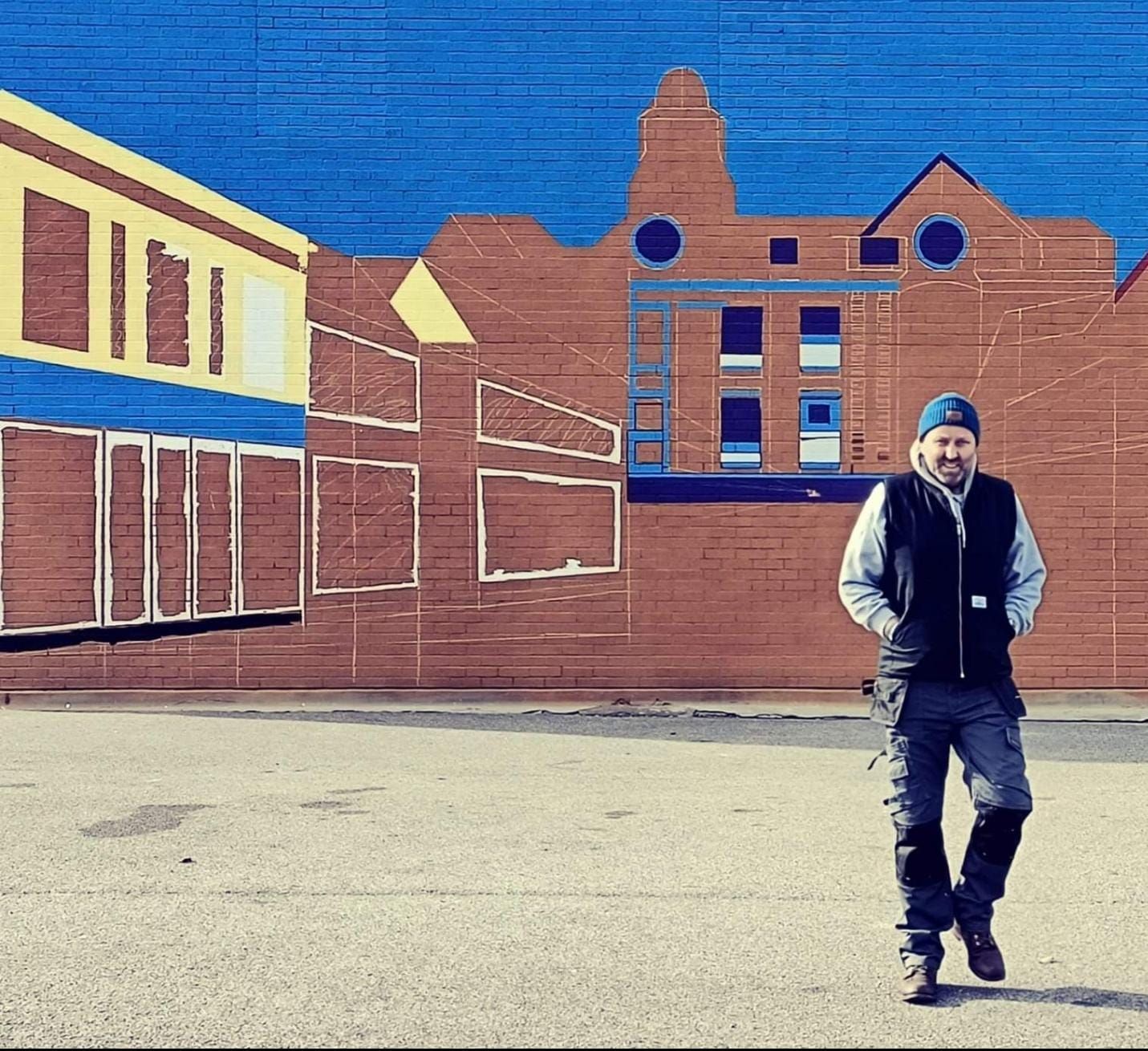 A man standing in front of a brick wall with buildings painted on it