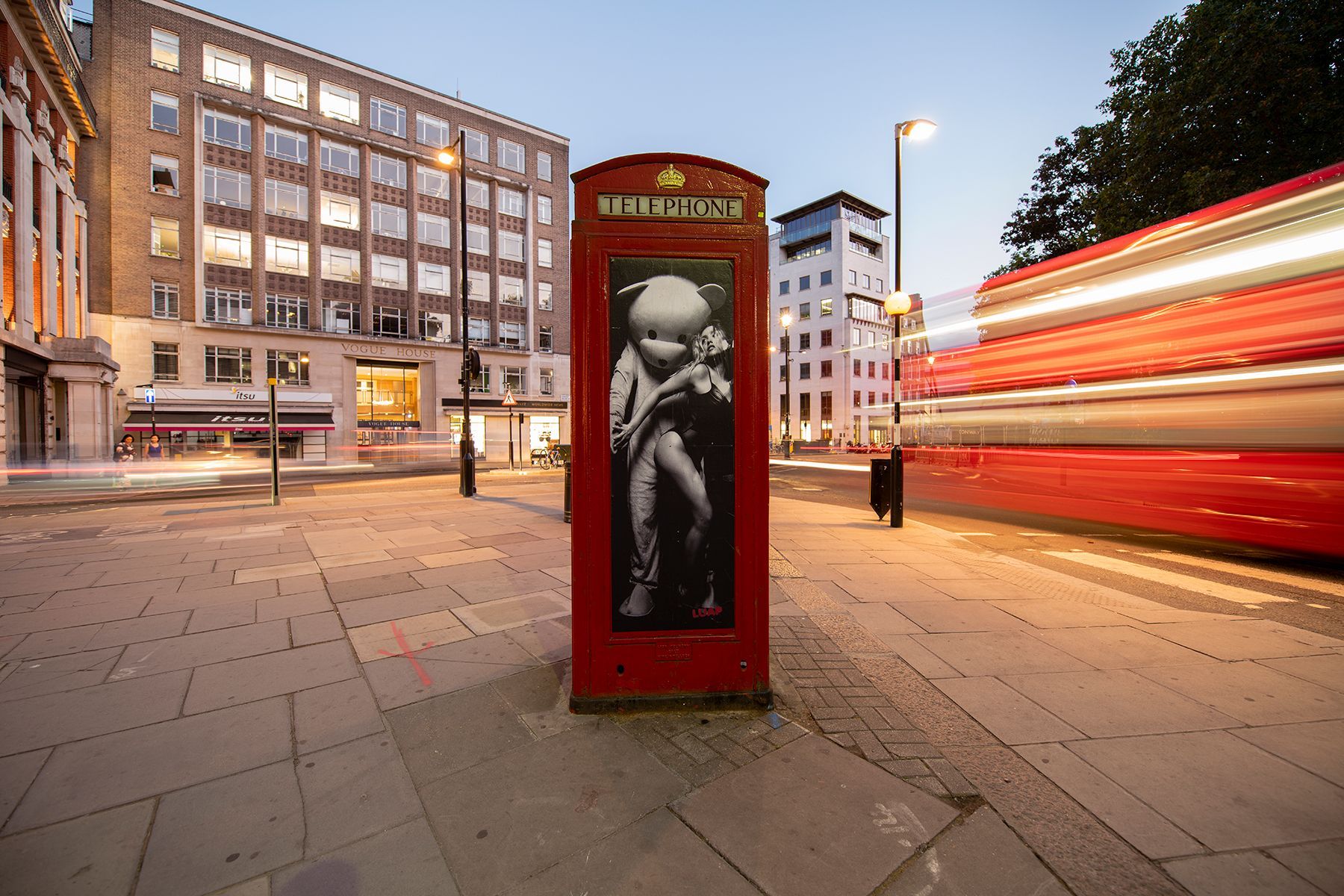 A red telephone booth with a picture of a baby on it is on the sidewalk in front of a bus.