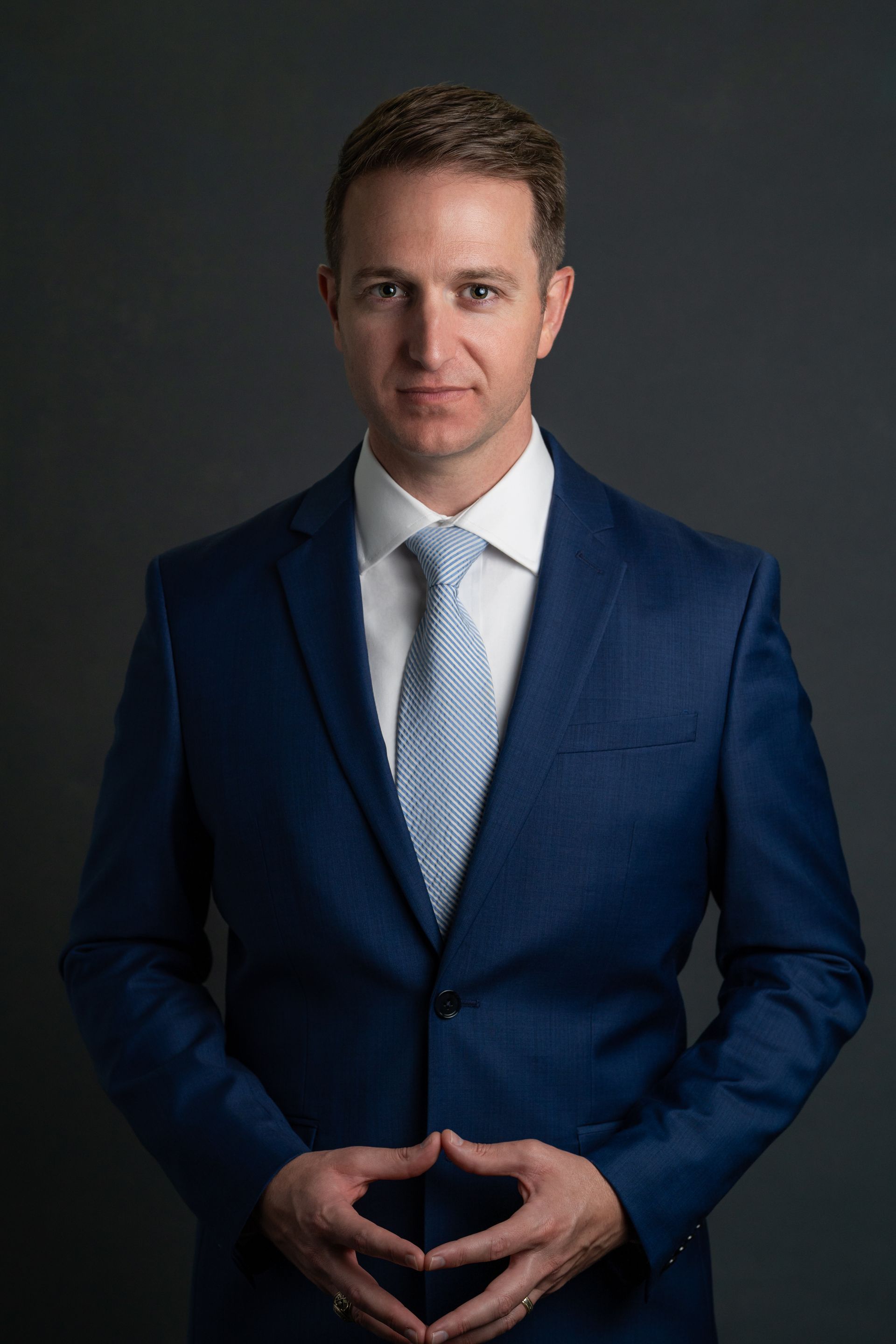 A man in a blue suit and tie is standing with his hands folded.