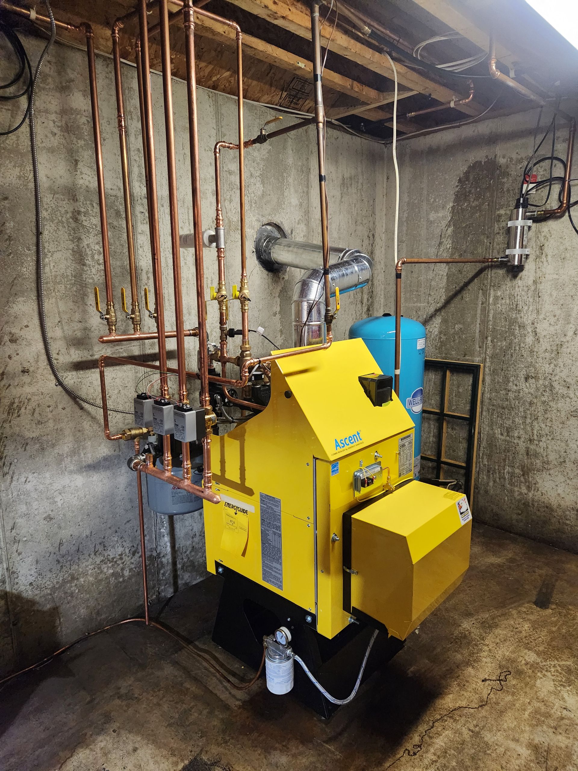 A yellow boiler is sitting in a room with copper pipes.