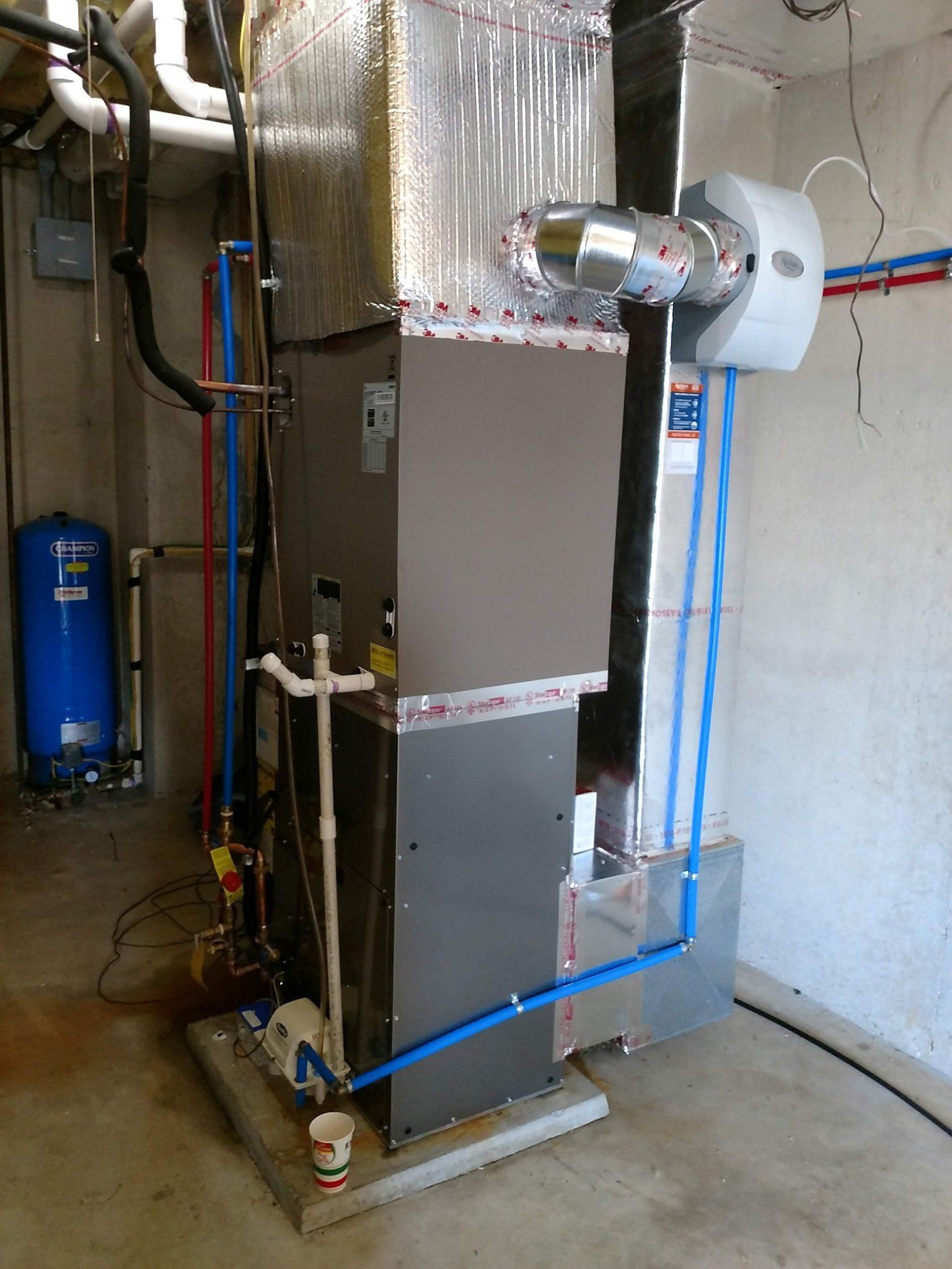 A large heater is sitting in a basement next to a blue tank.