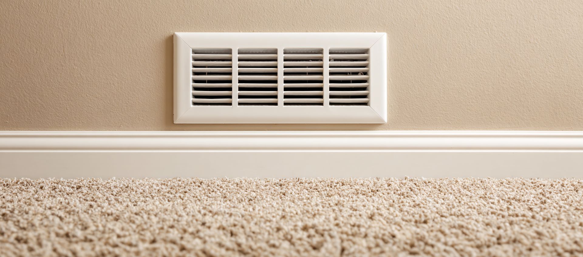 White air vent on a beige wall above white baseboard trim and beige carpet.