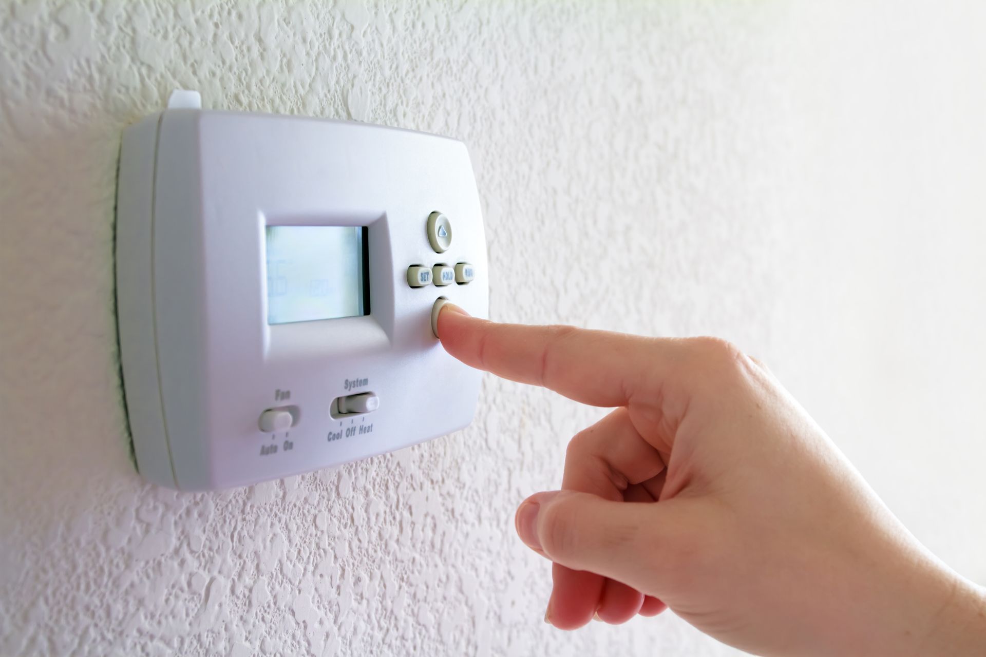 A person's finger presses a button on a white thermostat mounted on a white textured wall.