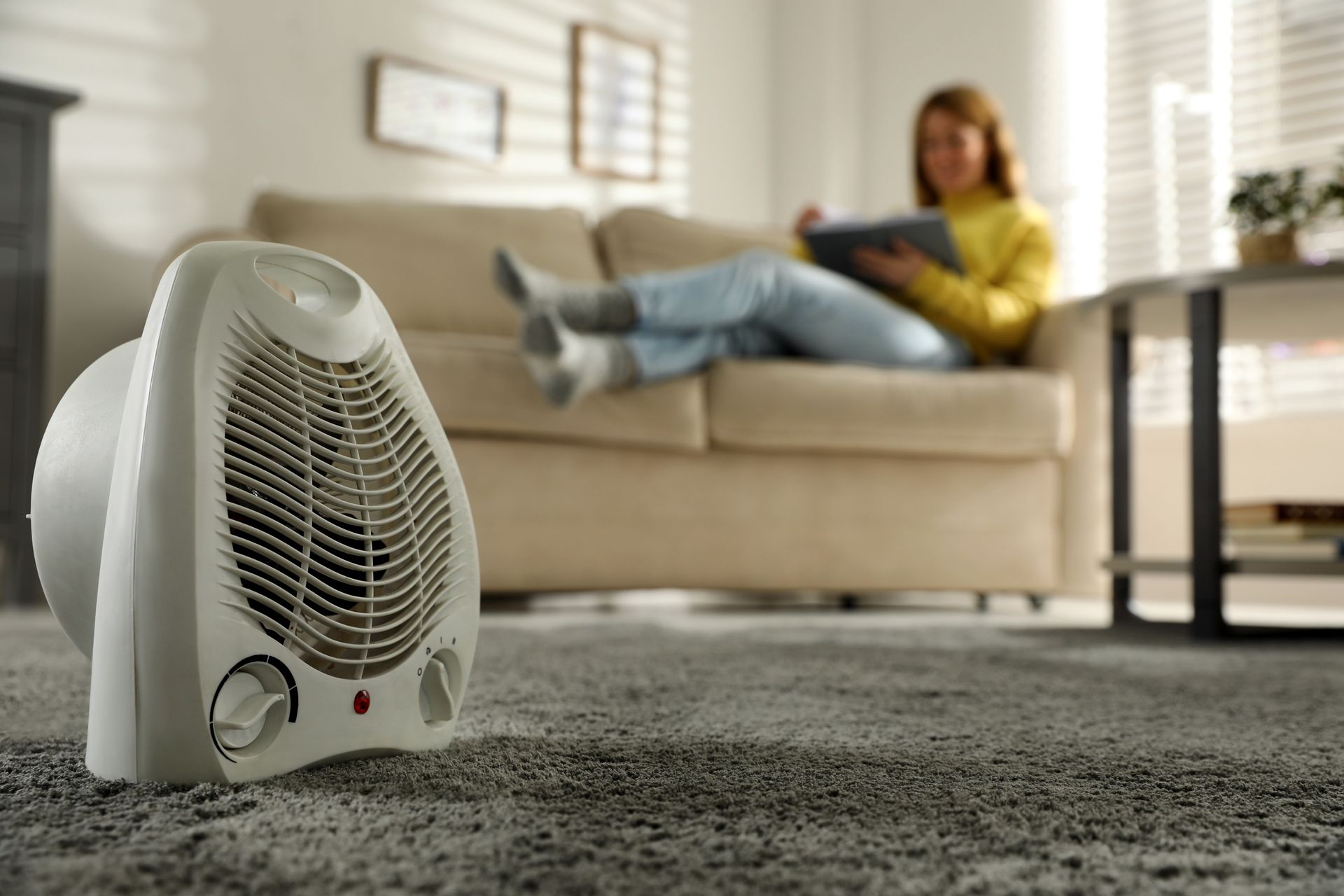 White space heater on a gray carpet in front of a couch, a person uses a tablet.