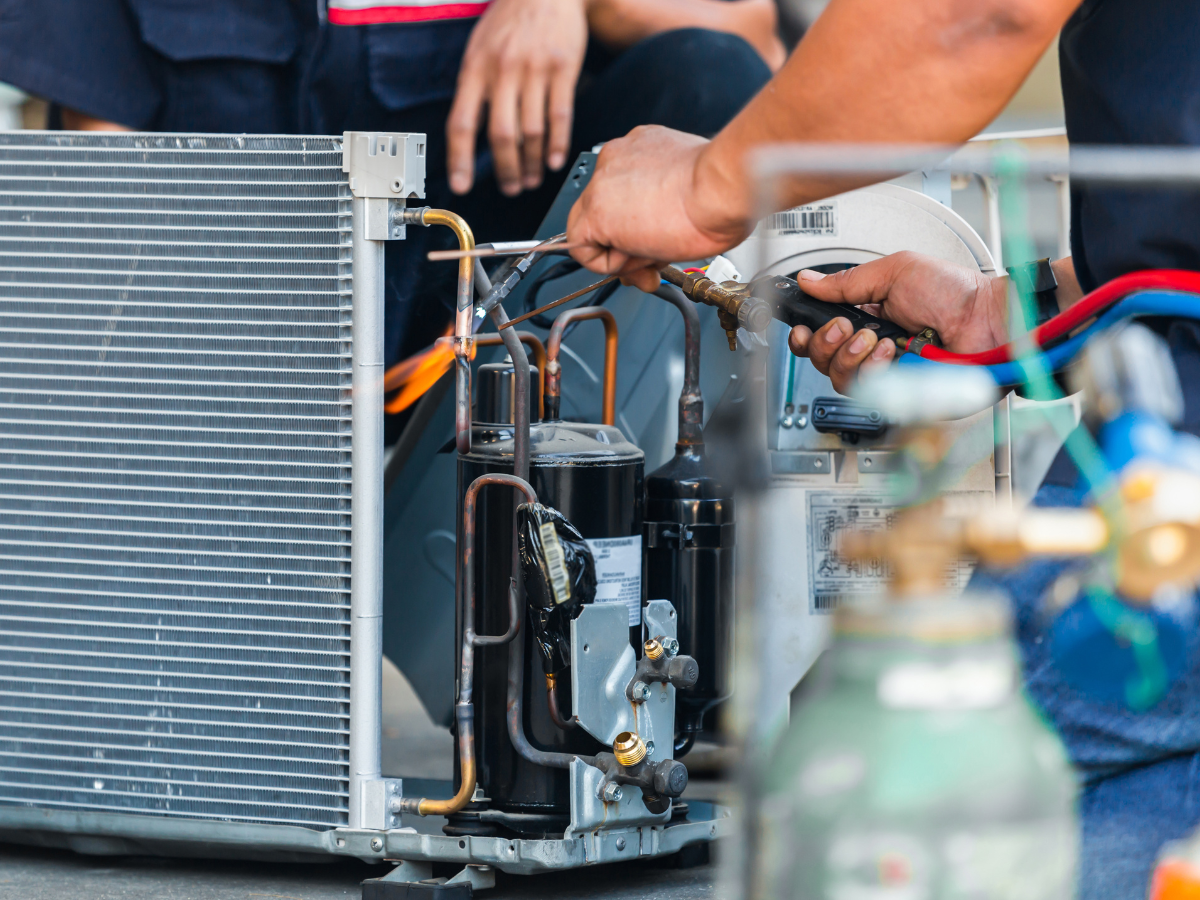 HVAC technician repairing an air conditioner with tools. Hands are visible, working near copper pipes and compressor.