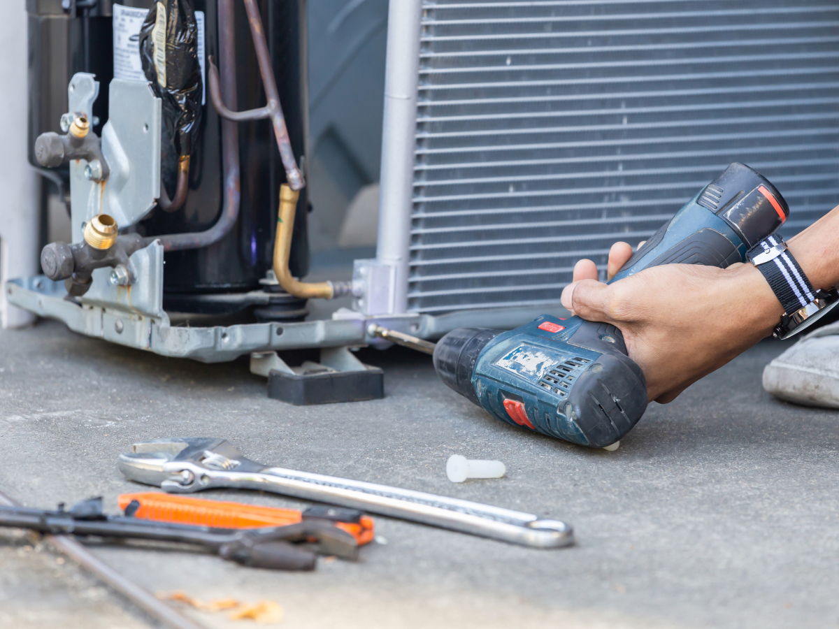 Person using a drill to repair an air conditioning unit outside. Tools lie on the concrete.