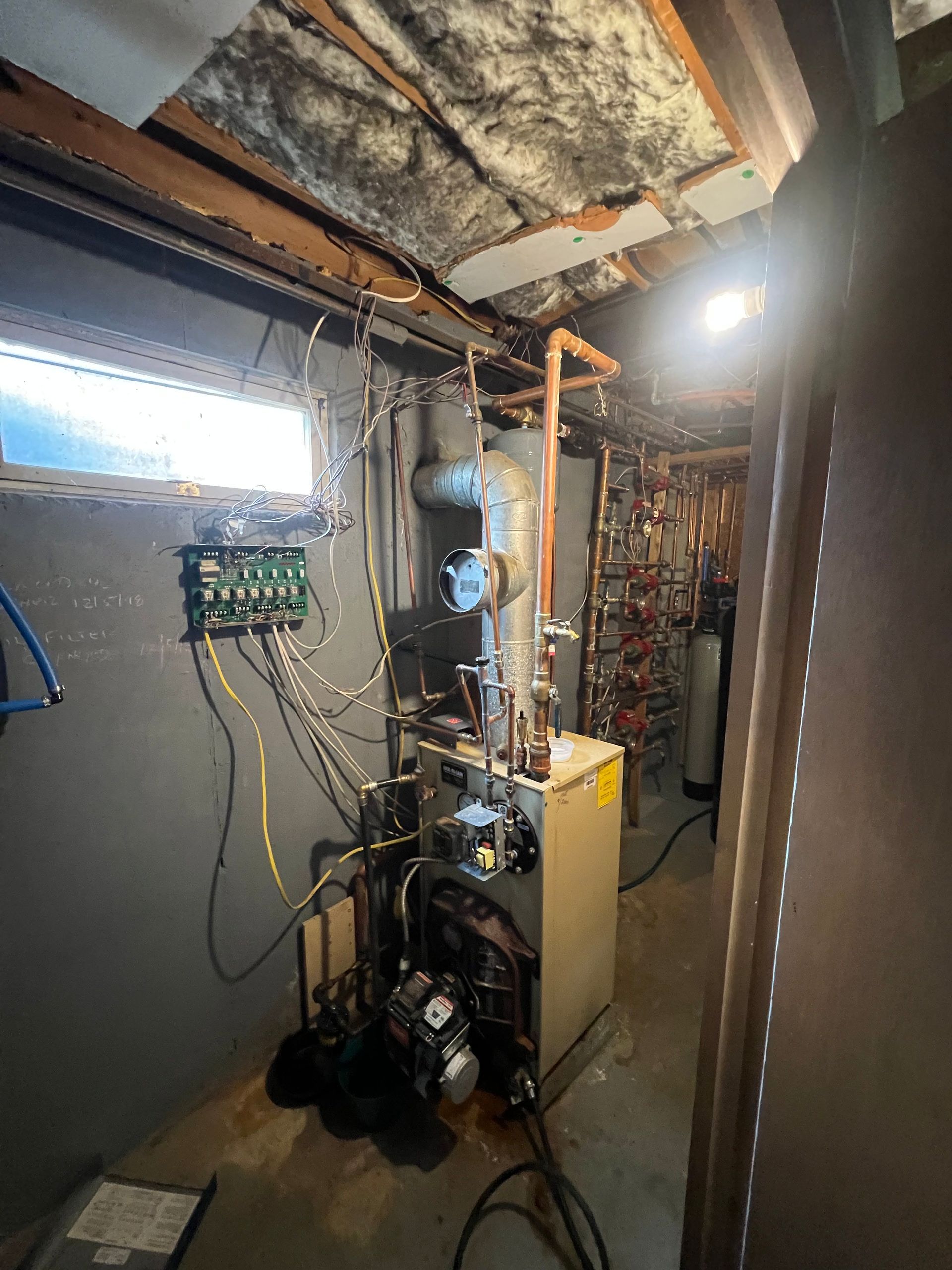 A boiler is sitting in a basement next to a window.