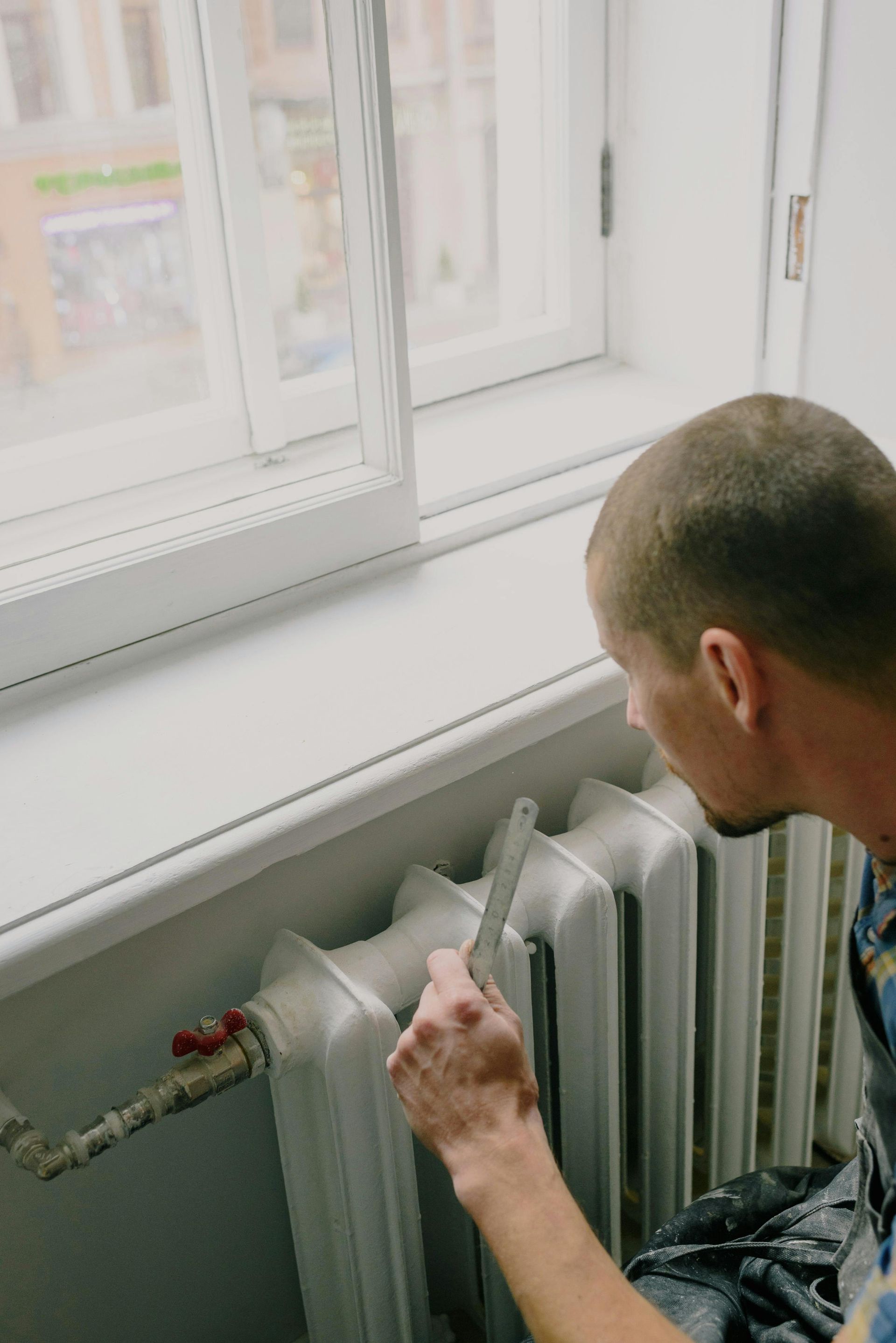 A man is sitting on a window sill fixing a radiator.