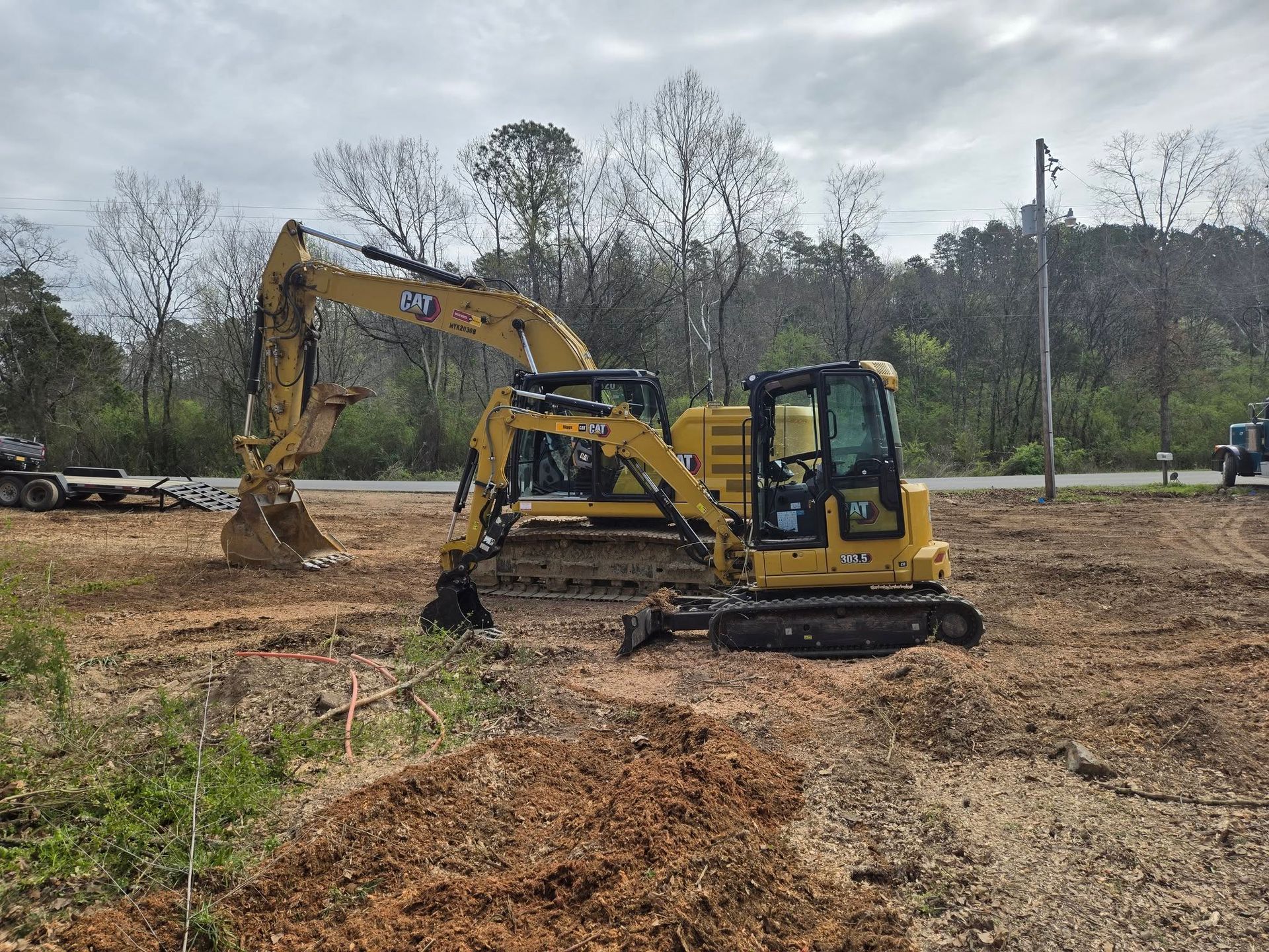 Two yellow excavators on a dirt lot, one digging amid trees and cloudy sky.