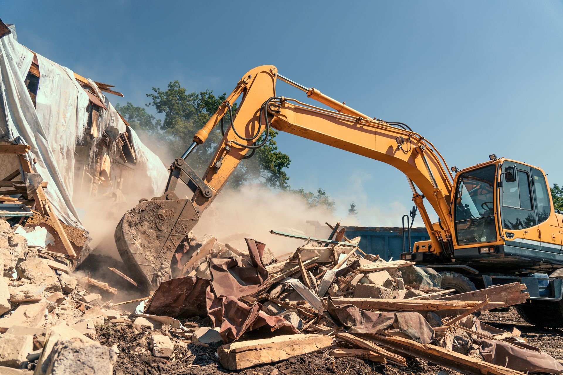 Yellow excavator demolishing a building, lifting debris amid dust and rubble under a blue sky