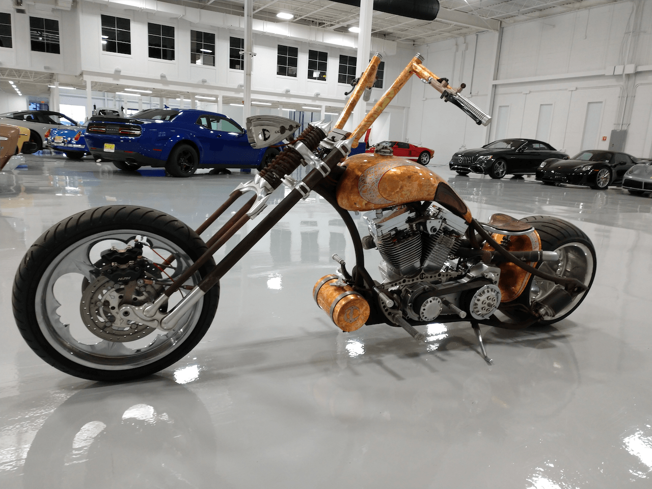 A chopper motorcycle is parked in a garage with cars parked in the background.