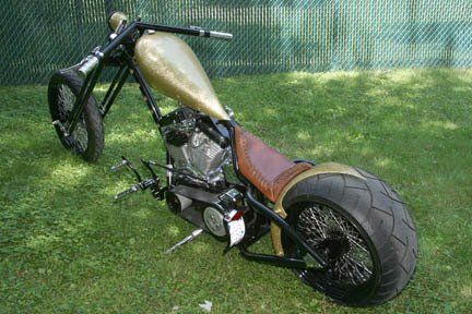 A chopper motorcycle is parked in the grass in front of a chain link fence.